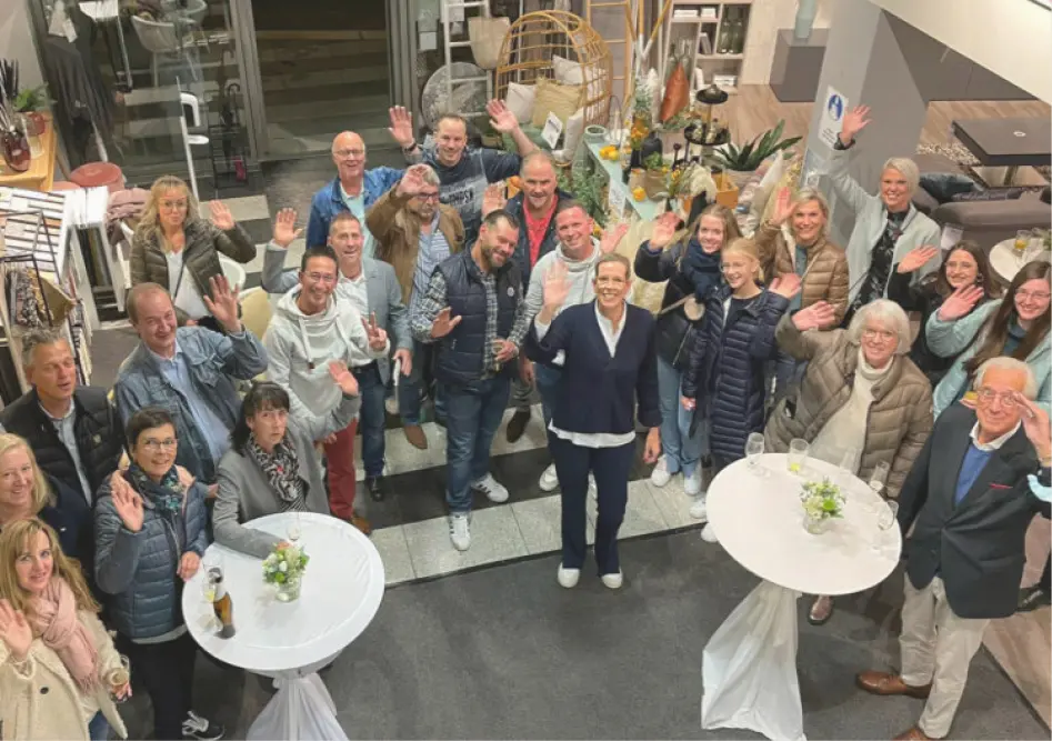 Mehrere Personen stehen im Möbelhaus um Stehtische und winken in die Kamera, Möbel und Dekoration im Hintergrund